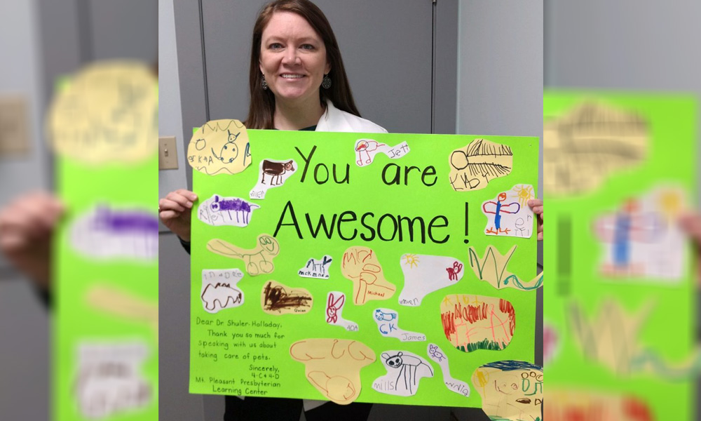 Woman smiling at the camera, holding a colorful handmade poster with children's drawings and the text "you are awesome!" in a veterinarian school setting.