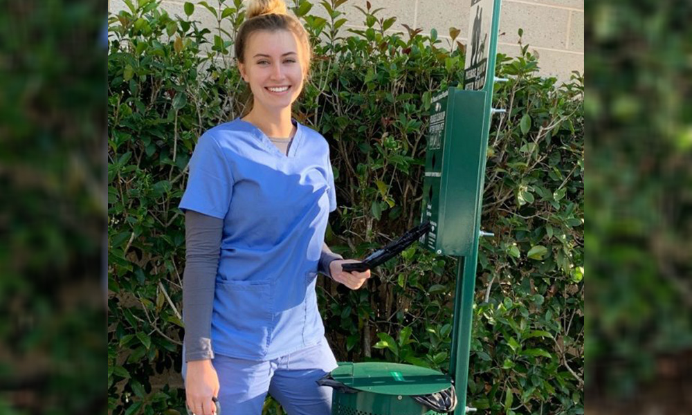 A smiling young veterinarian in blue scrubs stands beside a green mailbox, holding a clipboard in one hand and resting the other on her hip. Lush green bushes form the background.