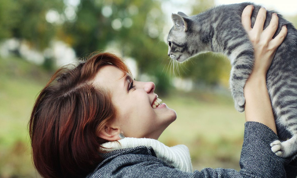 A joyful woman, perhaps a vet, with short brown hair holding and smiling at a gray tabby kitten outdoors, with trees in the soft-focus background.