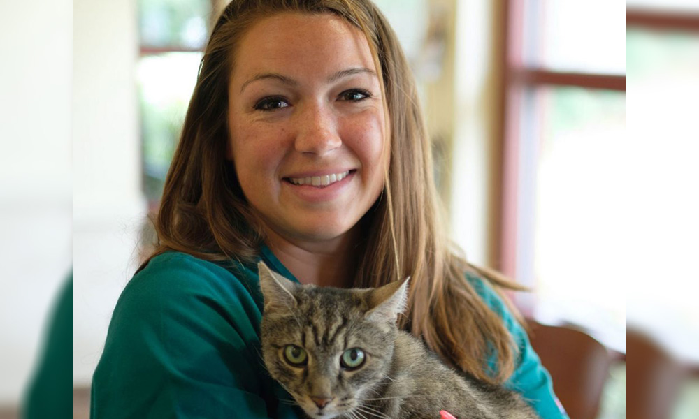 A smiling woman holding a tabby cat indoors at the vet, with a softly blurred background emphasizing their warm expressions.