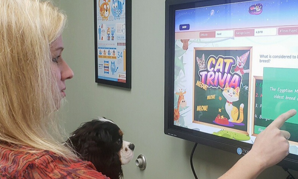 A woman and her dog interact with a computer screen displaying a "cat trivia" game at the veterinarian's office, pointing to a multiple-choice question about cat breeds.