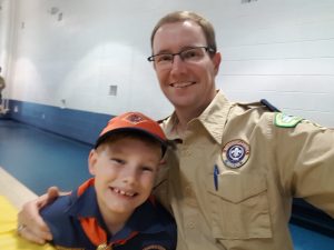 A smiling veterinarian in a scout leader uniform taking a selfie with a young boy in a scout hat and orange neckerchief in a gym.