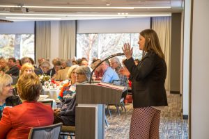 A veterinarian is presenting to an attentive audience seated at tables during a luncheon in a conference room with large windows that show leafy trees outside.