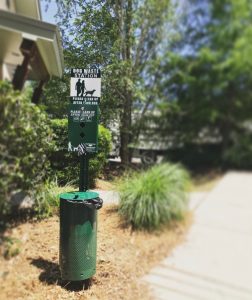 A pet waste station with a sign, bags, and a disposal bin beside a walking path bordered by shrubs under a sunny sky, endorsed by a local vet.