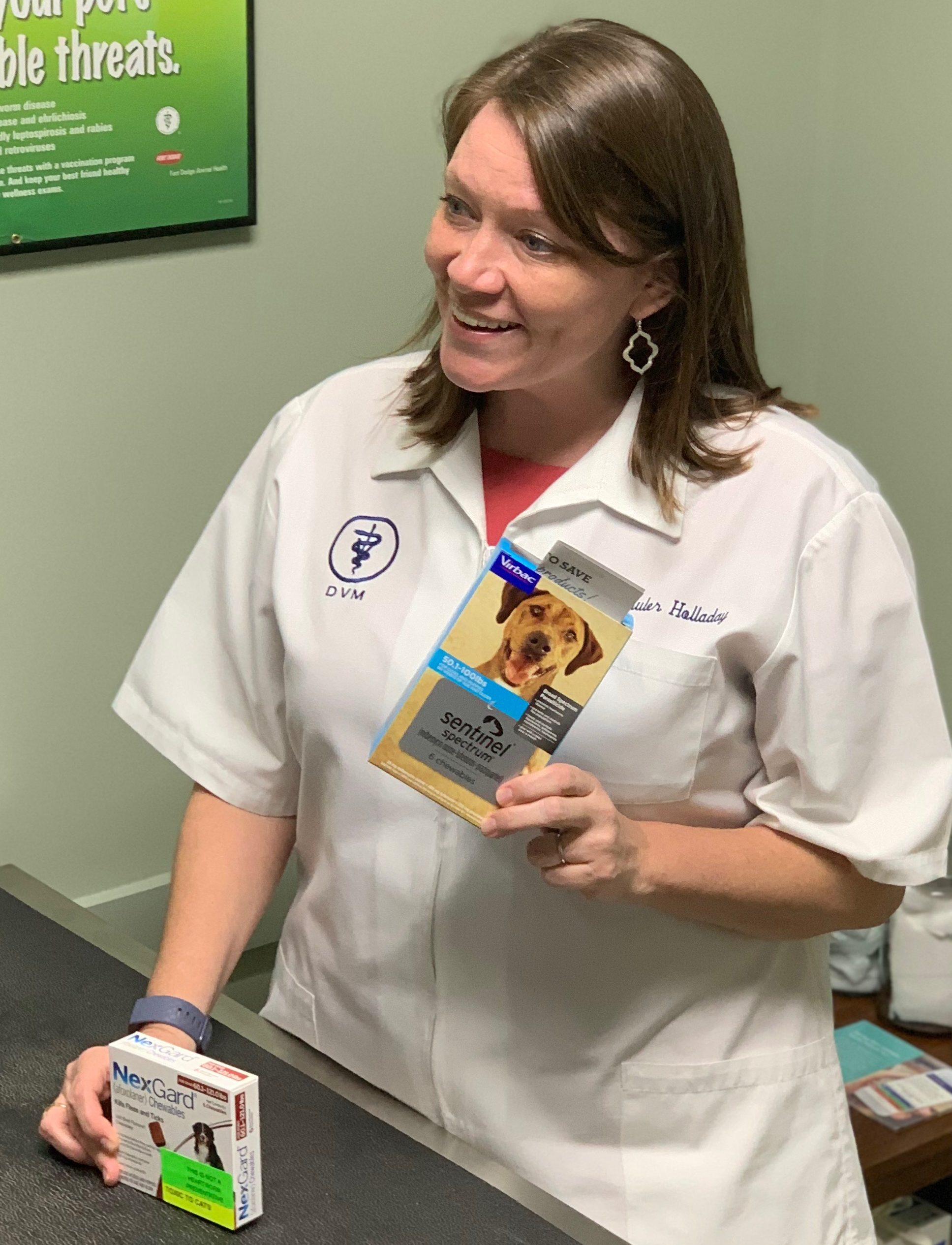 A smiling female vet in a white lab coat holding a box of pet medicine stands at a reception desk. Various veterinary products are visible on the counter.