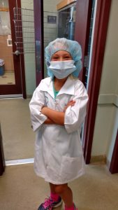 A young child dressed as a veterinarian in a lab coat, surgical mask, and cap stands smiling in a veterinary clinic, arms crossed, wearing bright pink sneakers.