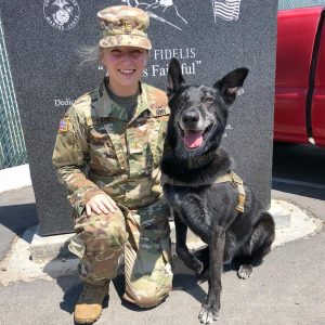 A female veterinarian in camouflage uniform smiling alongside a black military dog wearing a tactical vest, seated against a granite memorial with inscriptions.