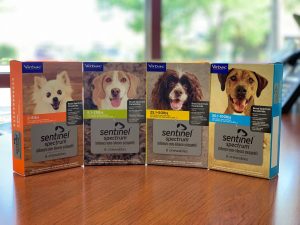 Four boxes of sentinel spectrum dog medication displayed on a vet's table, each featuring a different dog breed on the packaging, with a blurred background.