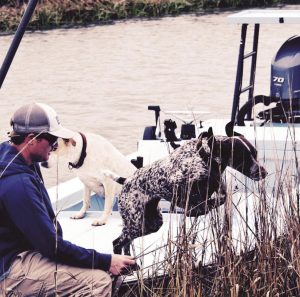 A man in a cap, who is a veterinarian, sits on a boat with three dogs, looking towards a river surrounded by tall grass. The boat's outboard motor is visible in the background.