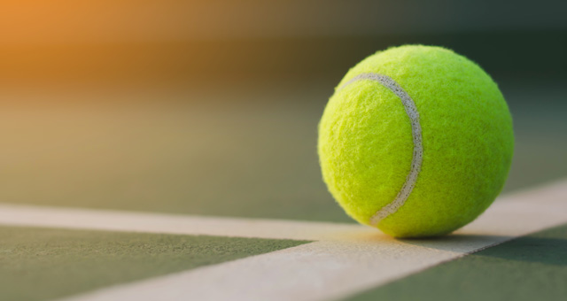 A close-up of a tennis ball on a court, catching sunlight, with its shadow casting slightly to the side. The ball rests on the white boundary line of the court.