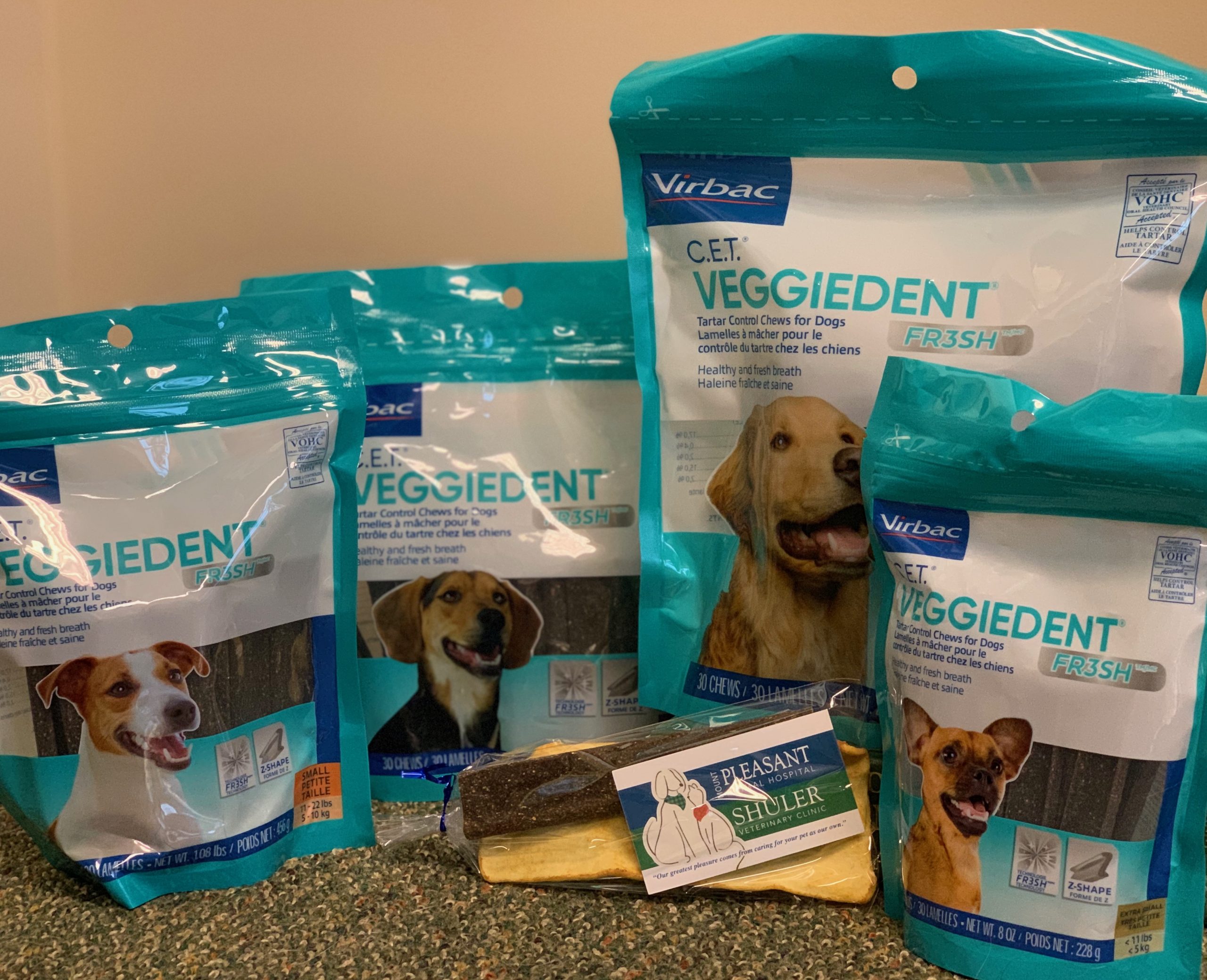 Three packages of Virbac Cet Veggiedent chews for dogs, recommended by veterinarians, featuring images of dogs on the labels, displayed on a counter with a rawhide bone in front.