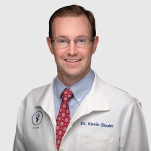 Professional portrait of a smiling male vet wearing glasses, a white lab coat with "Dr. Kevin Shuler, DVM" embroidered on it, and a red patterned tie.