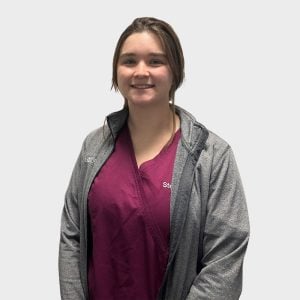 A young woman standing in front of a white background, with shoulder-length brown hair, wearing a burgundy top and a gray zip-up jacket with the name "Steph" embroidered on it, which identifies her as a veterinarian.